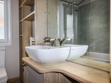 A bathroom with a white sink on a wooden countertop a mirror wooden shelves a window and a tiled shower at Beachcliff in Porth