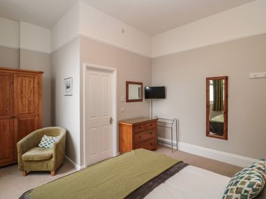 A bedroom with a bed a wooden wardrobe a green armchair a wooden chest of drawers a wall-mounted TV two mirrors and a door at Bay Tree House in Seaton Devon