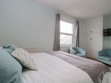 A bedroom with a bed a window with curtains two chairs a wall-mounted television and built-in cabinets at Bay Tree House in Seaton Devon