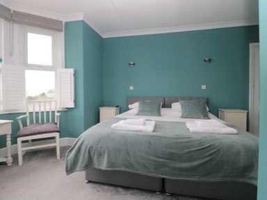A bedroom with teal walls a large bed with pillows and towels two bedside tables and a white chair next to a window at Bay Tree House in Seaton Devon