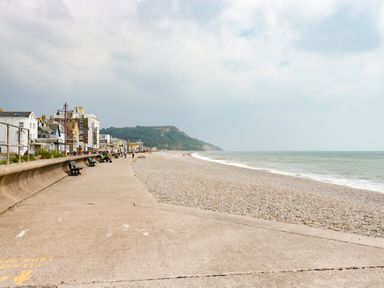 A pebble beach with a concrete promenade and benches next to houses and cliffs at Bay Tree House in Seaton Devon