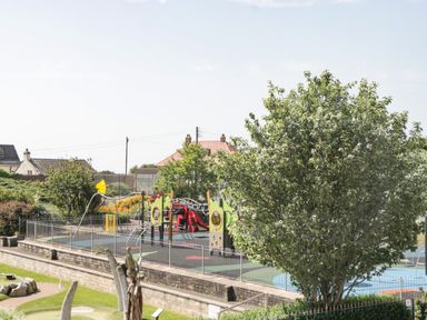 A fenced playground with climbing frames and slides behind a large tree in Seaton Devon