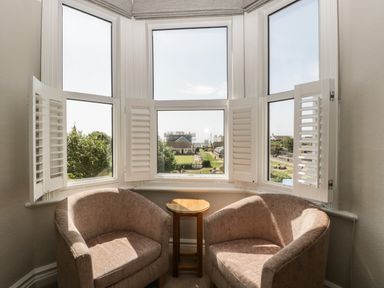 Two armchairs with a small round table between them by a bay window with white shutters at Bay Tree House in Seaton Devon