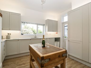 A kitchen with grey cabinets a wooden island table and a bottle of champagne with a glass on top at Bay Tree House in Seaton Devon