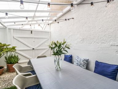 A covered outdoor dining area with a table and chairs a vase with flowers and cushions on the bench at Bay Tree House in Seaton Devon