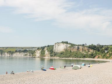 A pebble beach with tents and umbrellas by calm water and cliffs in the background at Bay Tree House in Seaton Devon