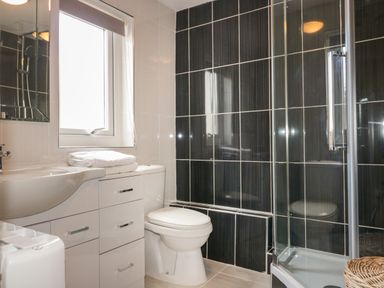 A bathroom with a toilet, a sink with drawers underneath, folded towels, a window, a shower with glass doors, and black tiled walls at Crantock Lodge in Crantock