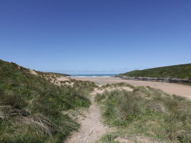 A sandy path through grassy dunes leading to a beach with hills on either side at Crantock Lodge in Crantock