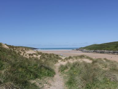 A sandy path through grassy dunes leading to a beach with hills on both sides at Crantock Lodge in Crantock