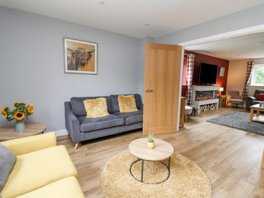 A living room with grey and yellow sofas a round wooden coffee table a painting of a bull and an open door showing another room with a fireplace and television at Talarddu Cottage in Builth Wells