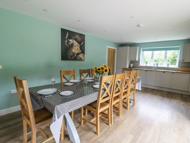 A dining area with a long table set with plates glasses and a vase of sunflowers next to a kitchen with cabinets and a window at Talarddu Cottage in Builth Wells