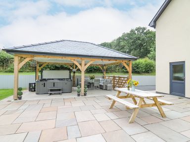 An outdoor patio area with a wooden picnic table and a covered gazebo with a hot tub and seating at Talarddu Cottage in Builth Wells