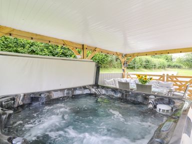 An outdoor covered area with a bubbling hot tub and a seating area with a glass table and chairs with sunflowers on the table at Talarddu Cottage in Builth Wells