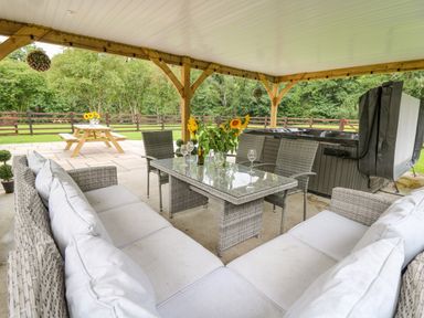 A covered outdoor seating area with a glass table surrounded by chairs and couches next to a hot tub and picnic table in a garden at Talarddu Cottage in Builth Wells