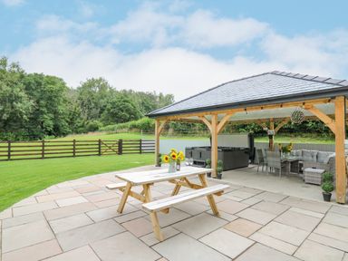An outdoor patio with a wooden picnic table flowers on the table a covered seating area with sofas and a hot tub at Talarddu Cottage in Builth Wells