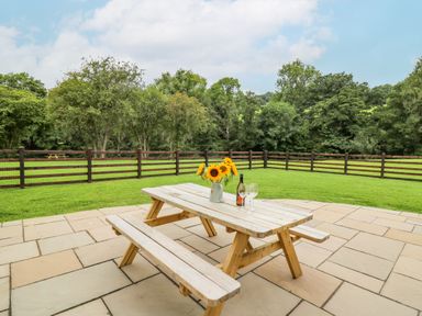 A wooden picnic table with sunflowers in a vase and wine with two glasses on a stone patio with a fenced grass lawn and trees at Talarddu Cottage in Builth Wells