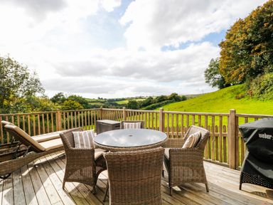 An outdoor wooden deck with a round table surrounded by wicker chairs and a lounge chair with green fields and trees in the background at Frogwell Chapel in Frogwell near Callington