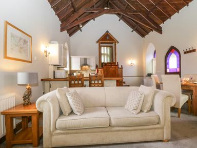 A living room with a beige sofa and wooden furniture including a side table with a lamp and a dining table near stained glass windows at Frogwell Chapel in Frogwell near Callington