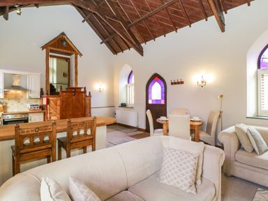 An open living space with a kitchen, wooden bar stools, a beige sofa, dining table with chairs, and stained glass windows at Frogwell Chapel in Frogwell near Callington