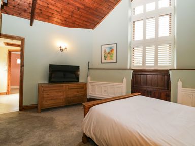 A bedroom with a wooden bed frame white bedding wooden dresser with a TV wall sconce and tall windows with white shutters at Frogwell Chapel in Frogwell near Callington