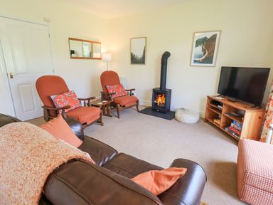 A living room with two armchairs a wood stove a television on a wooden stand and a leather sofa at Limhus Cottage in Keswick