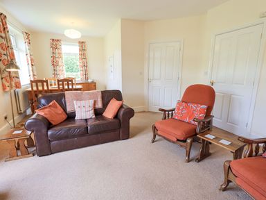 A living room with a brown leather sofa orange cushioned chairs a wooden dining table and floral curtains at Limhus Cottage in Keswick