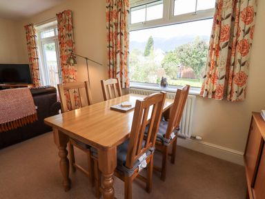 A dining table with four chairs next to a large window with floral curtains in a living room at Limhus Cottage in Keswick