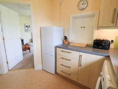 A kitchen area with a white refrigerator wooden cabinets with handles a countertop knives in a block a cutting board a black toaster a wall clock and a washing machine at Limhus Cottage Keswick