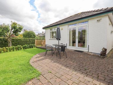 A patio with table chairs and an umbrella outside a white building with a garden and a tree at Limhus Cottage in Keswick