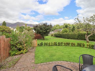 A garden with green grass shrubs a tree and outdoor chairs on a paved patio at Limhus Cottage in Keswick
