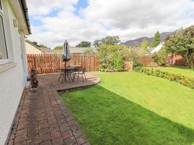 A backyard with a paved patio area with table and chairs a wooden fence and grassy lawn at Limhus Cottage in Keswick