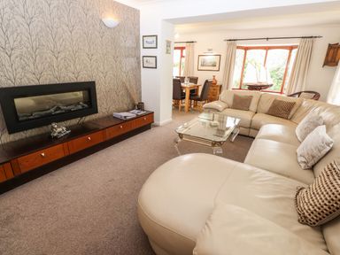 A living room with a beige sectional sofa a glass coffee table a mounted fireplace and a dining area at Fir Trees in Bowness-On-Windermere