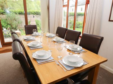 A dining room with a wooden table set with white plates bowls forks knives wine glasses and black chairs at Fir Trees in Bowness-On-Windermere