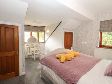 A bedroom with a bed covered by a purple blanket and yellow towels a white dressing table with a chair near windows at Fir Trees in Bowness-On-Windermere
