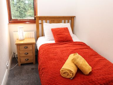 A single bed with red bedding and yellow towels on top next to a wooden nightstand with a lamp by a window at Fir Trees in Bowness-On-Windermere