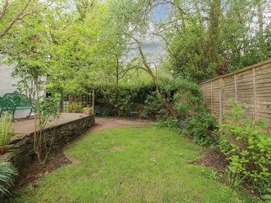A garden with grass a stone patio green metal chairs and trees surrounded by a wooden fence at Fir Trees in Bowness-On-Windermere