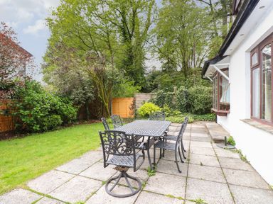 An outdoor patio with a metal table and chairs next to a grassy garden with trees and shrubs at Fir Trees in Bowness-On-Windermere