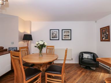 A dining area with a round wooden table and four chairs with black seats a vase of flowers on the table a black armchair with a pillow two framed pictures and a painting on the walls at The Sanctuary in Windermere