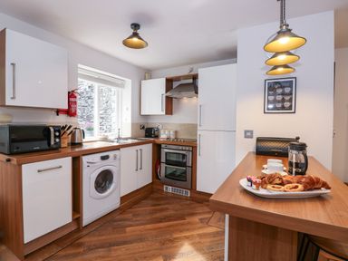 A kitchen with wooden floors white cabinets a washing machine an oven a microwave and a counter with pastries and coffee at The Sanctuary in Windermere