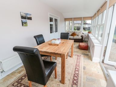 A dining area with a wooden table and black chairs next to a sitting area with a sofa and windows at Deanrise in Shilbottle near Alnwick