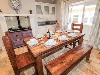 A dining room with a wooden table set with plates bowls wine glasses and a bottle of wine next to a stove and white cabinets at Deanrise in Shilbottle near Alnwick