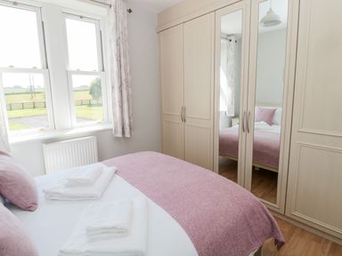 A bedroom with a bed having pink pillows and blanket white towels on the bed a large wardrobe with mirrored doors and windows with curtains at Deanrise Shilbottle near Alnwick