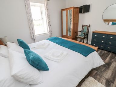 A bedroom with a bed with white linens and teal pillows a wooden wardrobe a chair and a dark blue dresser with a round mirror at Deanrise in Shilbottle near Alnwick