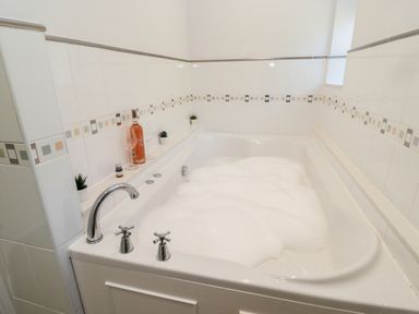 A bathroom with a white bathtub filled with bubbles bottles and glasses on the ledge at Deanrise in Shilbottle near Alnwick