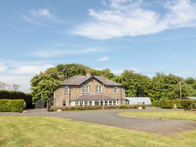 A stone house with a driveway and green lawn surrounded by trees at Deanrise in Shilbottle near Alnwick