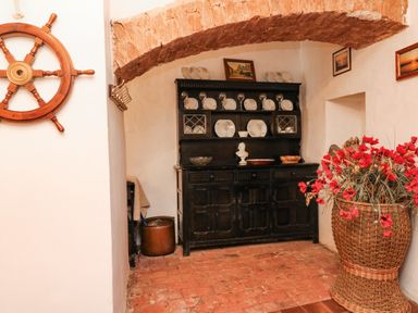 A room with a wooden ship wheel on the wall a black cupboard with plates and glasses and a wicker basket with red flowers at Harbour Cottage in Watchet