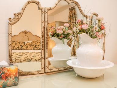A white ceramic pitcher with flowers inside placed in a matching bowl on a table in front of a decorative mirror reflecting a bed with floral bedding and curtains in a bedroom
