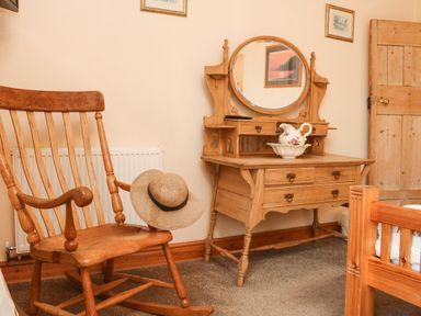 A wooden rocking chair with a straw hat next to a wooden vanity dresser with a round mirror and a ceramic pitcher in a bedroom at Harbour Cottage in Watchet