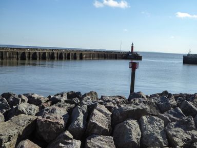A harbor with large rocks in the foreground a breakwater with a red lighthouse and calm water at Harbour Cottage in Watchet