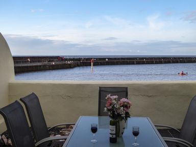 An outdoor seating area with a glass table and chairs overlooking a harbor with people walking on the pier and a person kayaking on the water at Harbour Cottage in Watchet